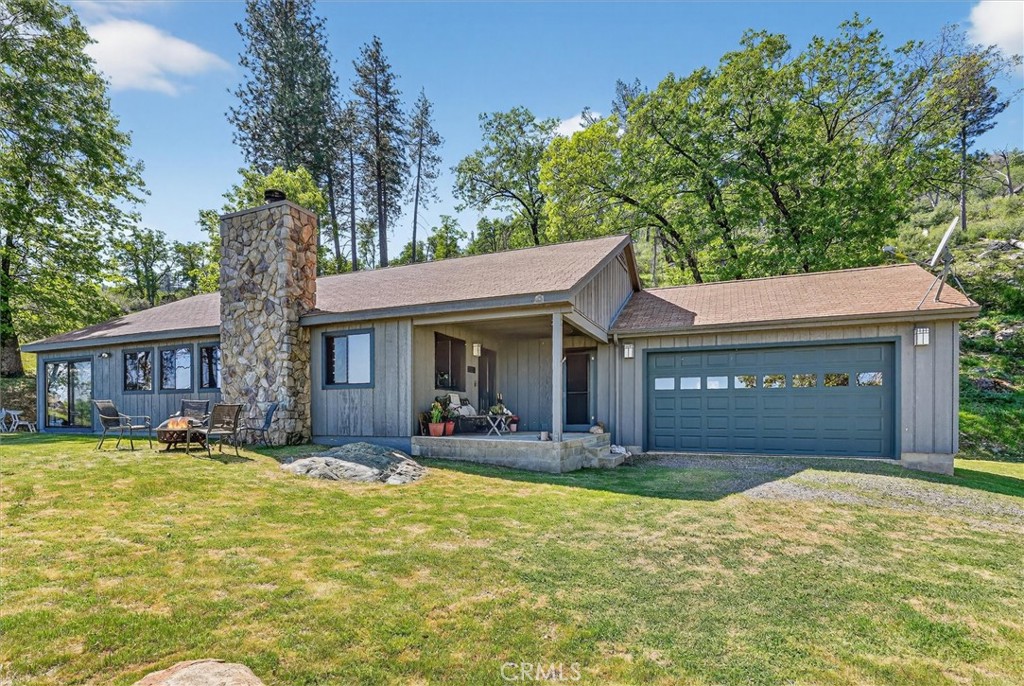 Gray single-story home with a stone chimney, covered entry, and attached garage among mature trees.