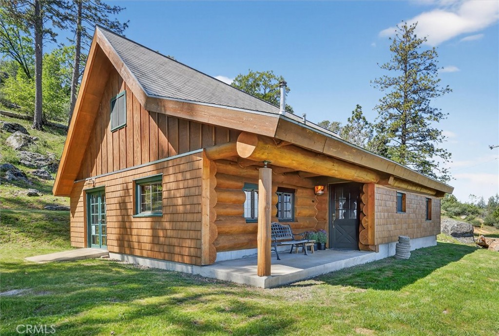 Rustic log cabin with a covered porch and bench on a grassy, pine-covered hillside.