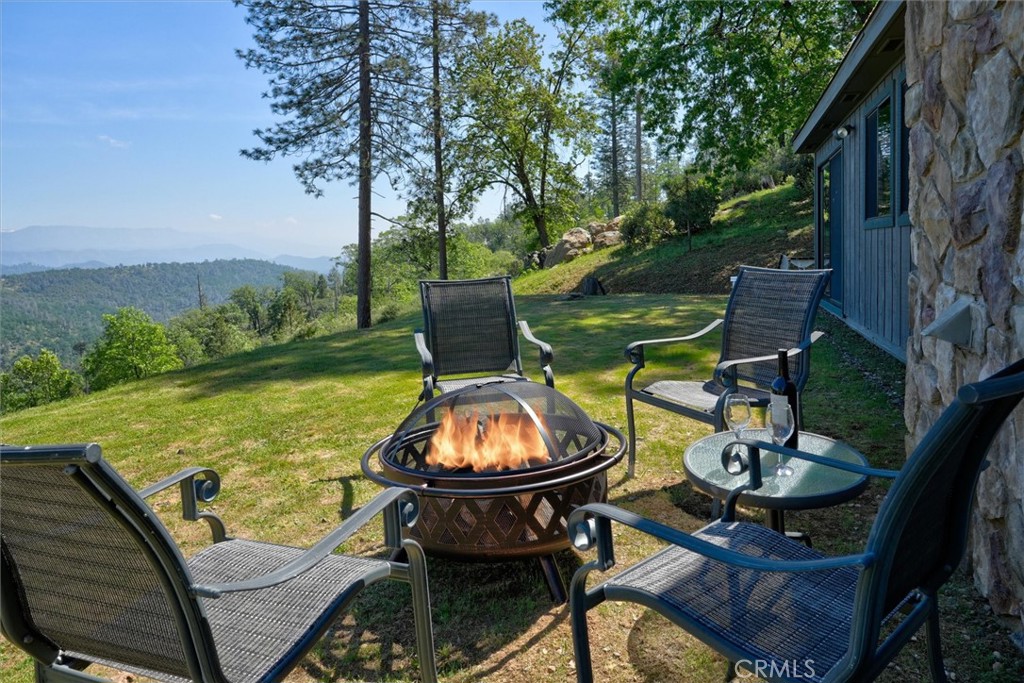 Outdoor chairs surround a lit fire pit beside the home, overlooking wooded hills and distant mountains.