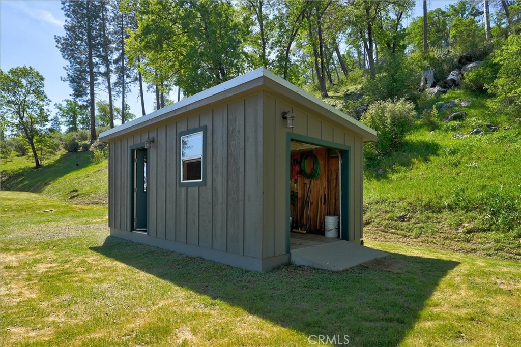 Detached gray-green storage shed with its door open on a grassy, wooded hillside.