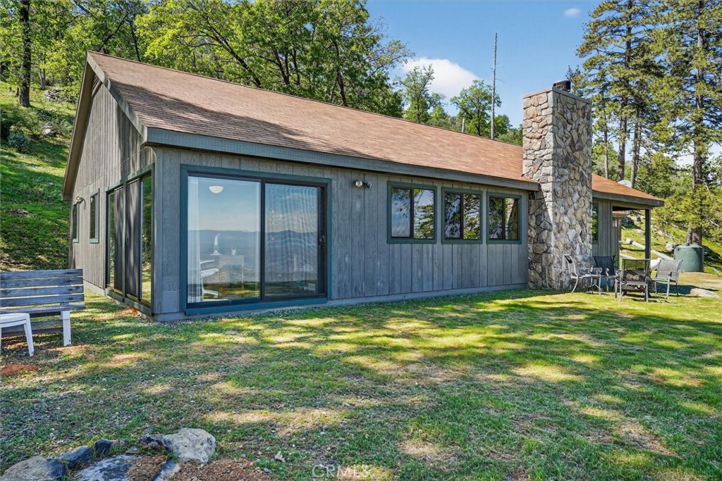 Blue-gray home with large sliding glass doors and a tall stone chimney beside a grassy, wooded yard.