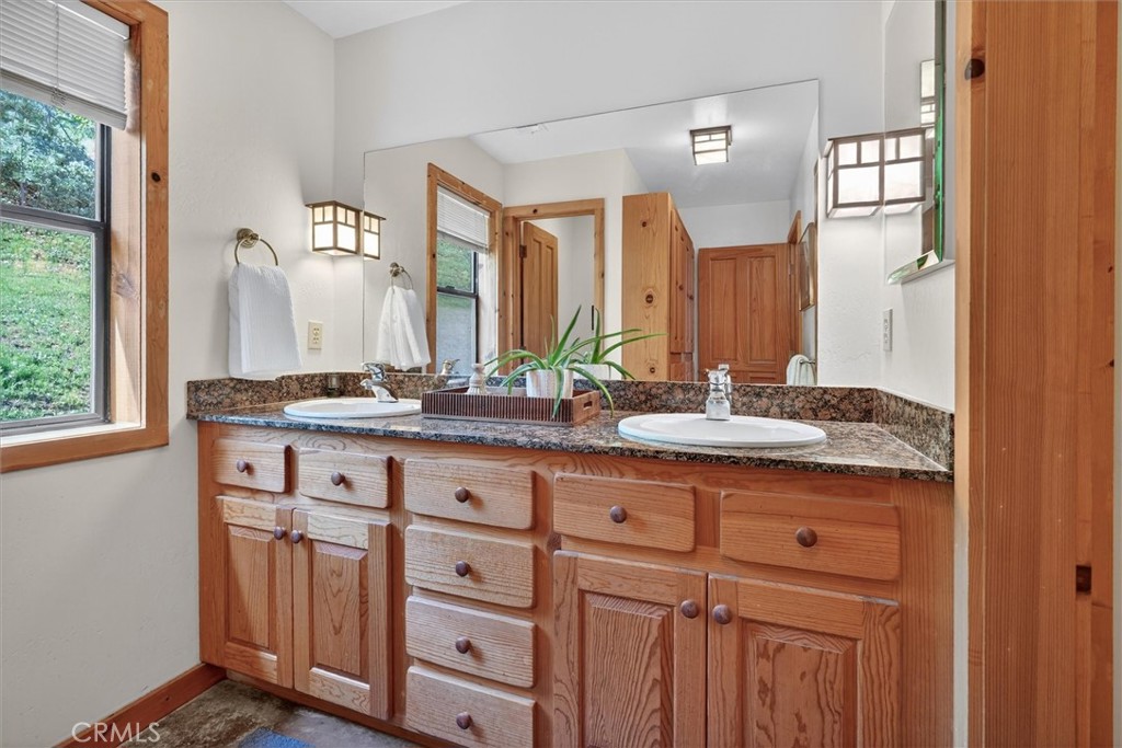 Bathroom vanity with two sinks, warm wood cabinetry, and a window overlooking greenery.