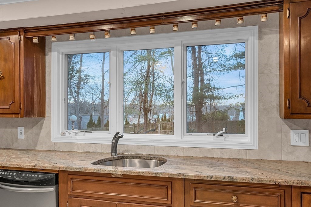 Kitchen sink beneath a three-pane window overlooking water and trees, framed by wood cabinets.