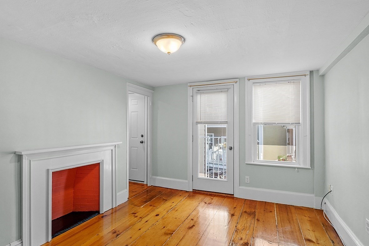 Unfurnished living room with a white-trimmed fireplace, hardwood floors, and a glass door beside a window.