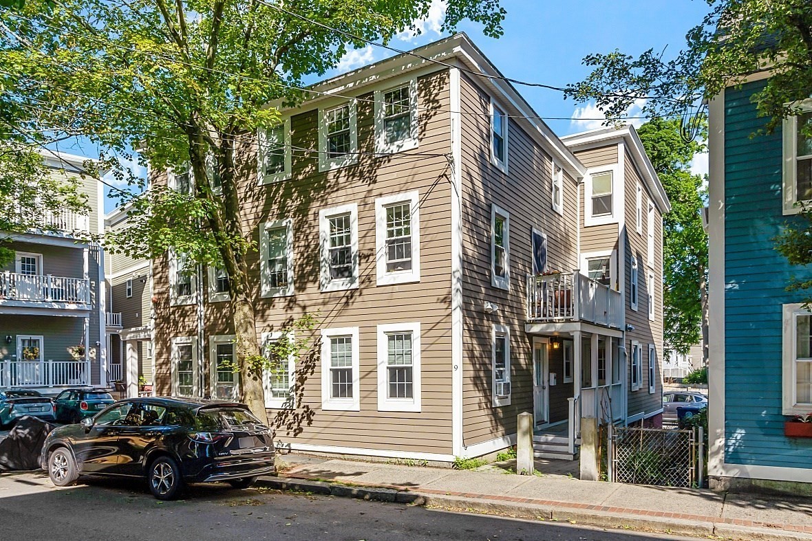 Multi-story beige-sided residence with white trim, a small upper balcony, and shaded street frontage.
