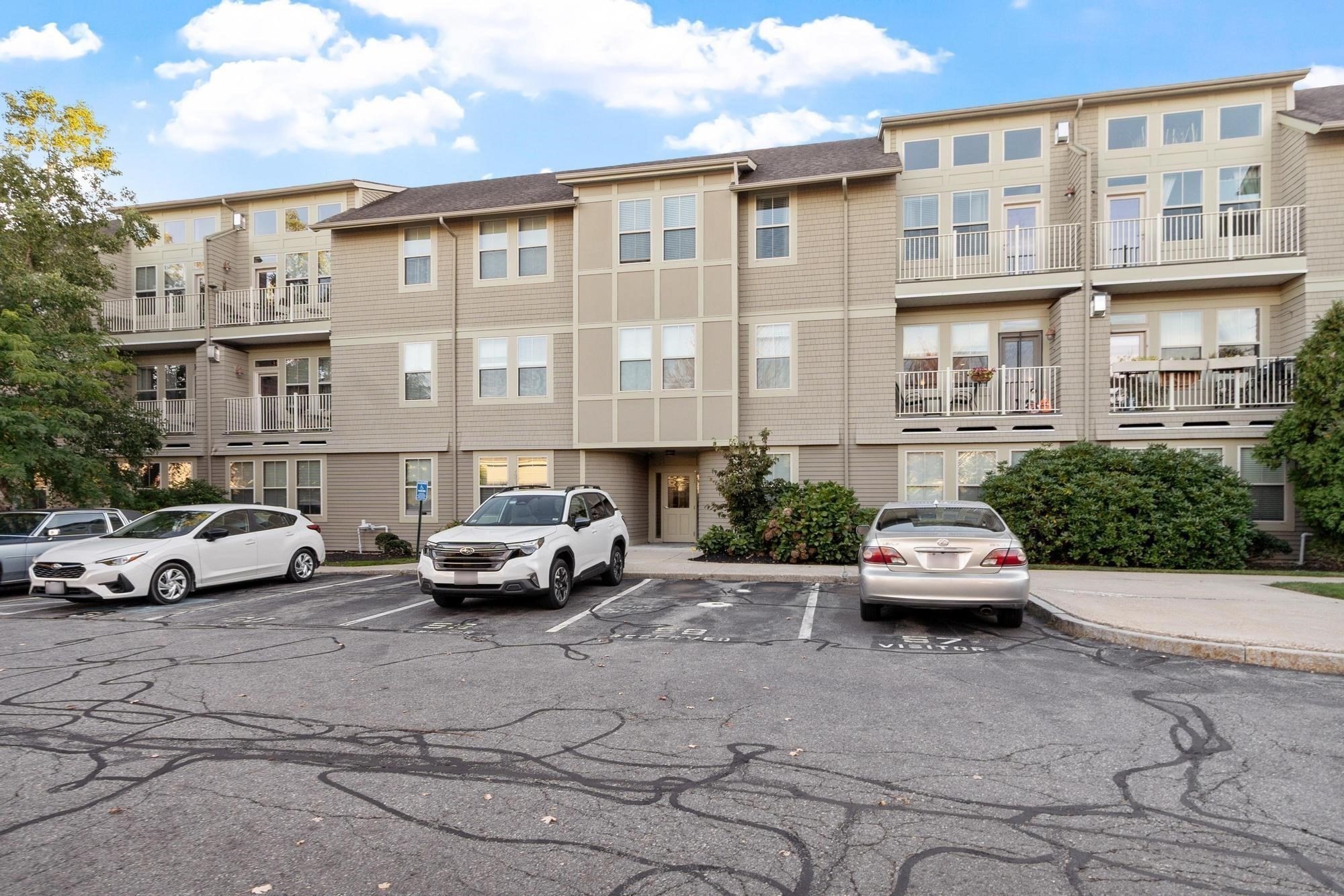 Multi-story beige condominium building with white-railed balconies, landscaping, and a parking lot in front.