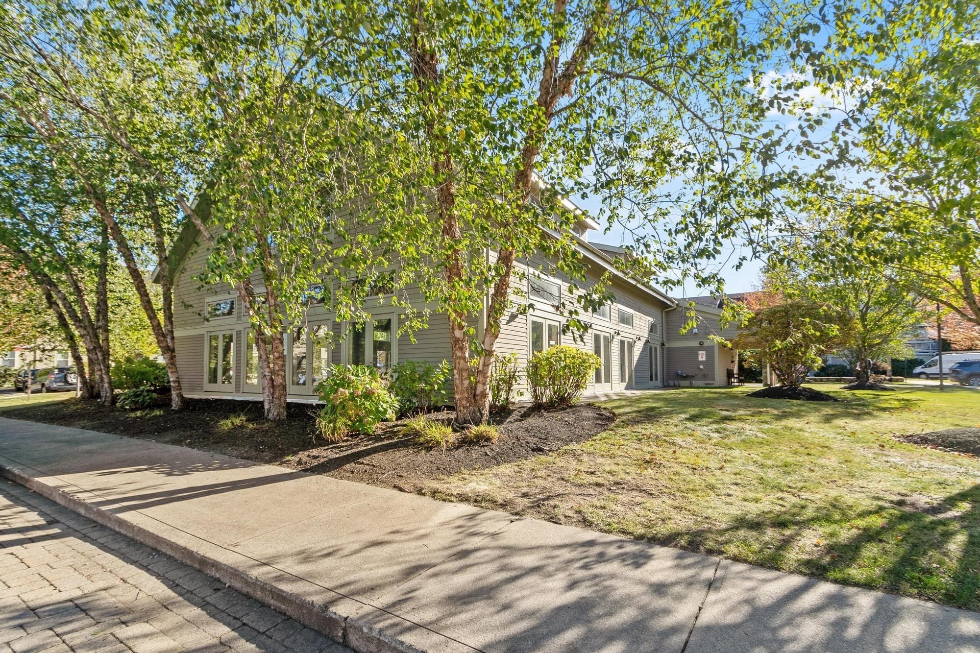 Gray-sided residential building partly screened by mature trees, with landscaped beds and a front lawn.