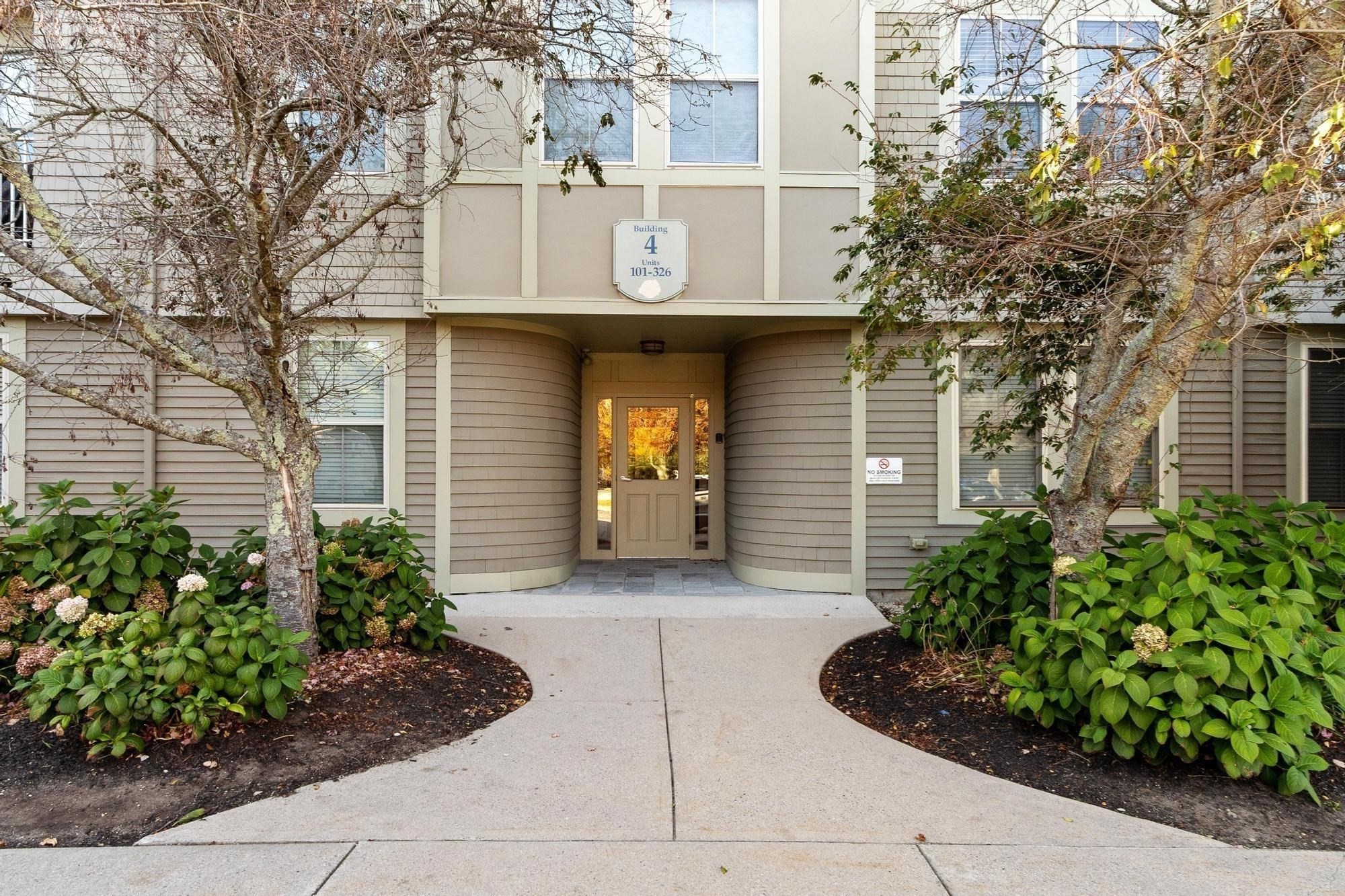 Residential building entrance with a recessed door, Building 4 sign, landscaped shrubs, and mature trees.