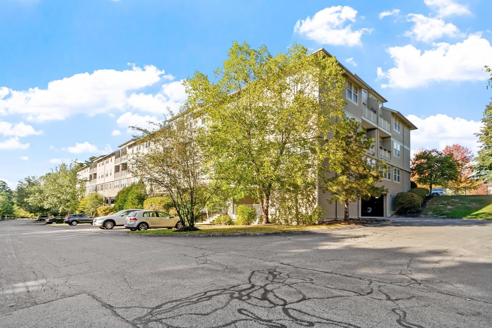 Multi-story beige condominium building with balconies behind leafy trees and a parking lot.