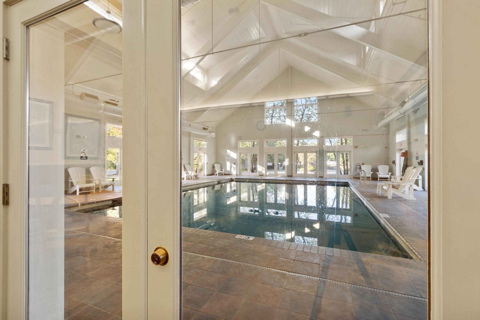 Indoor pool with a tiled deck, white lounge chairs, and a high vaulted ceiling.