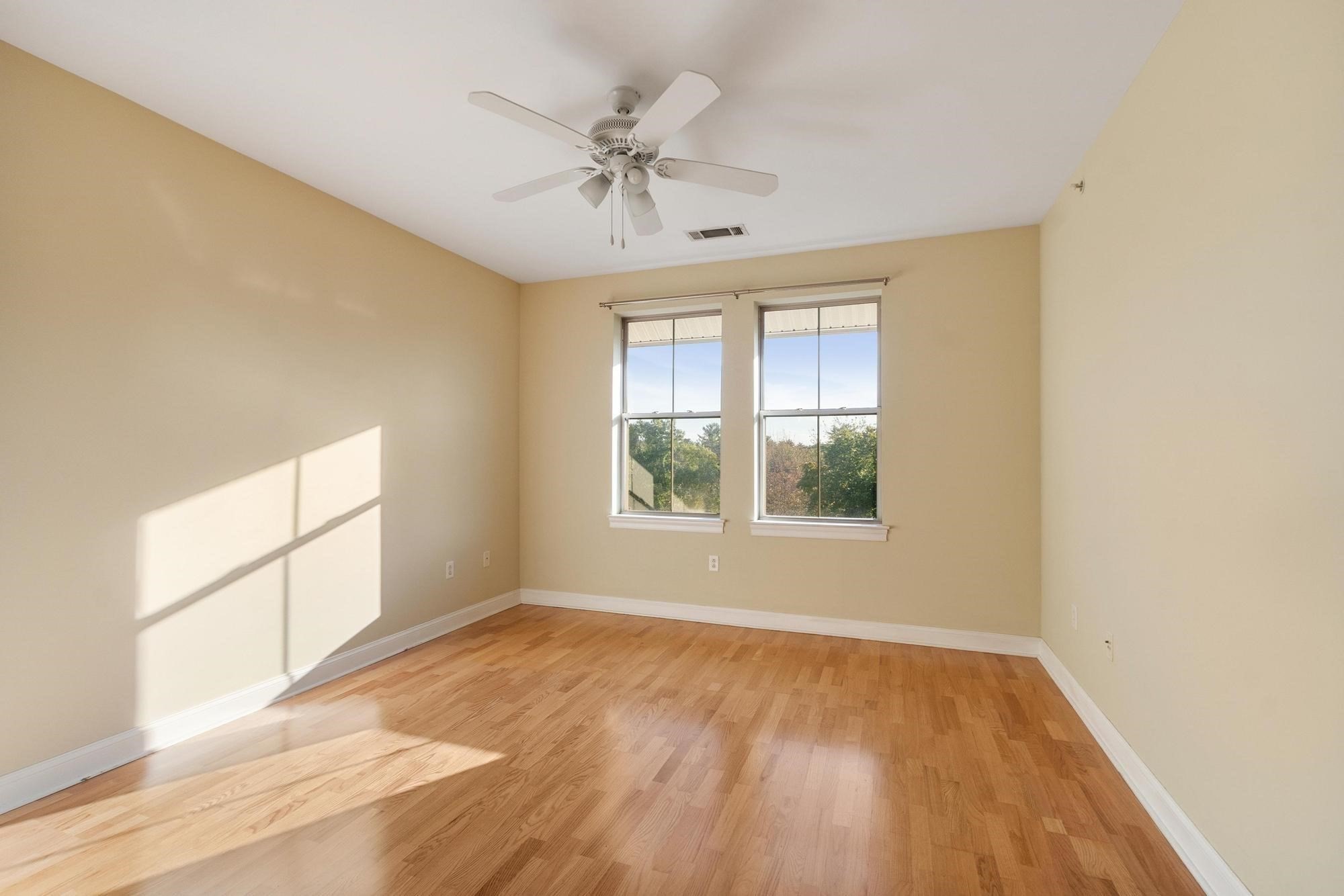 Empty room with hardwood floors, two windows, and a ceiling fan.