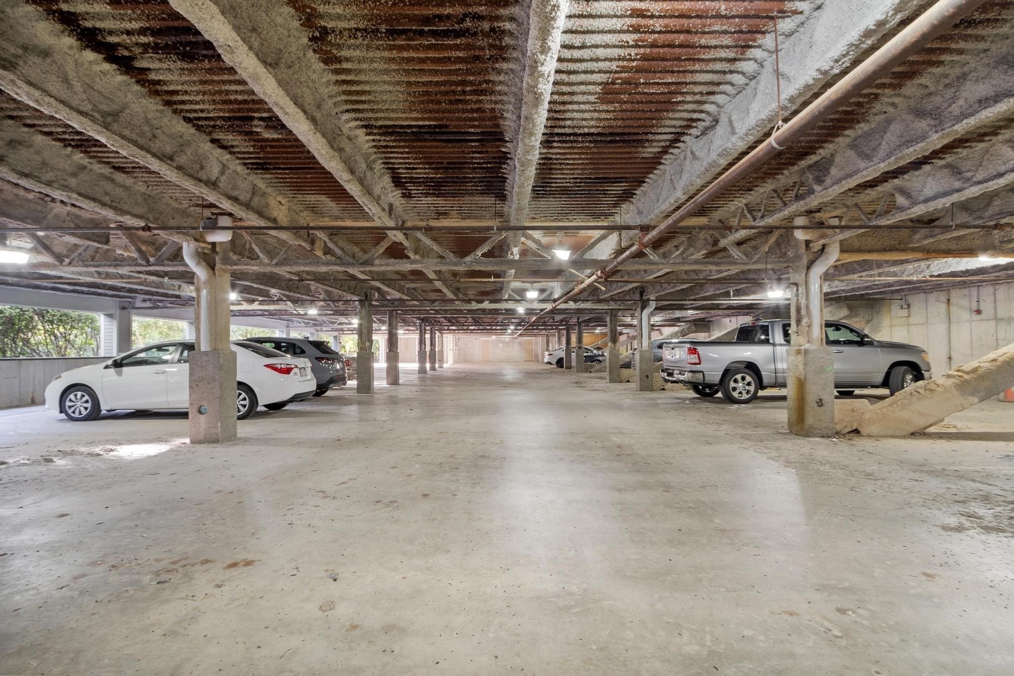 Shared parking garage with exposed concrete beams, overhead pipes, and parked cars.