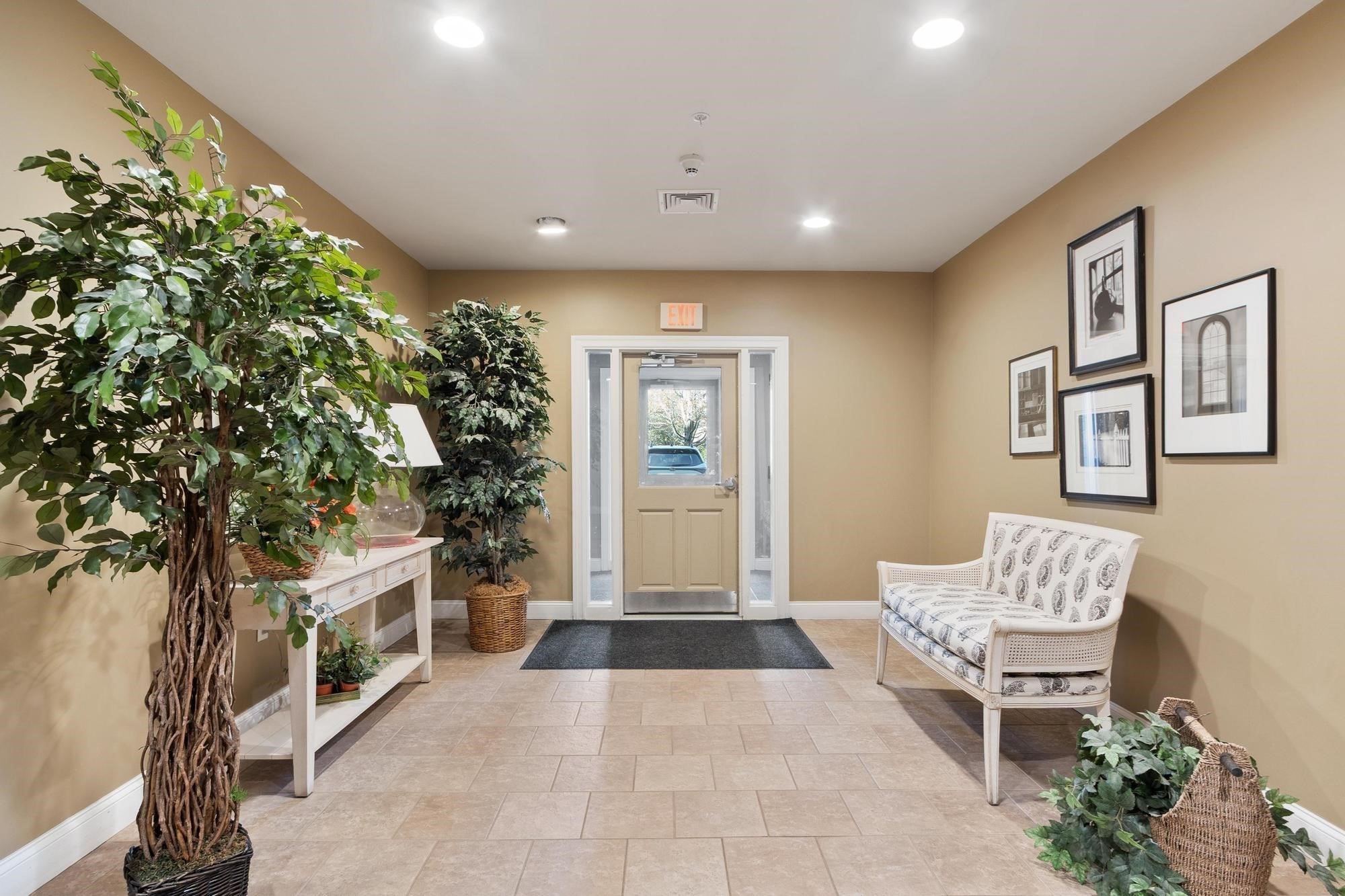 Furnished entry lobby with a glass-paneled door, potted trees, framed artwork, and a white loveseat.