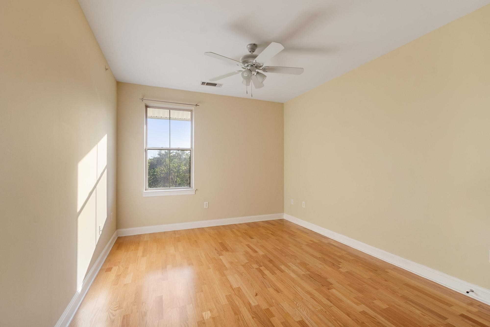 Empty room with hardwood floors, a ceiling fan, and sunlight streaming through a window.