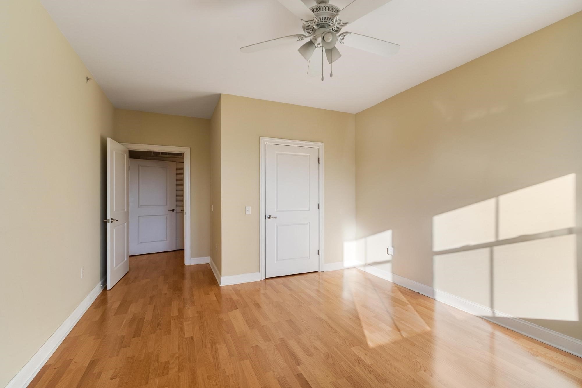 Empty room with hardwood floors, a white ceiling fan, and sunlight across the beige walls.