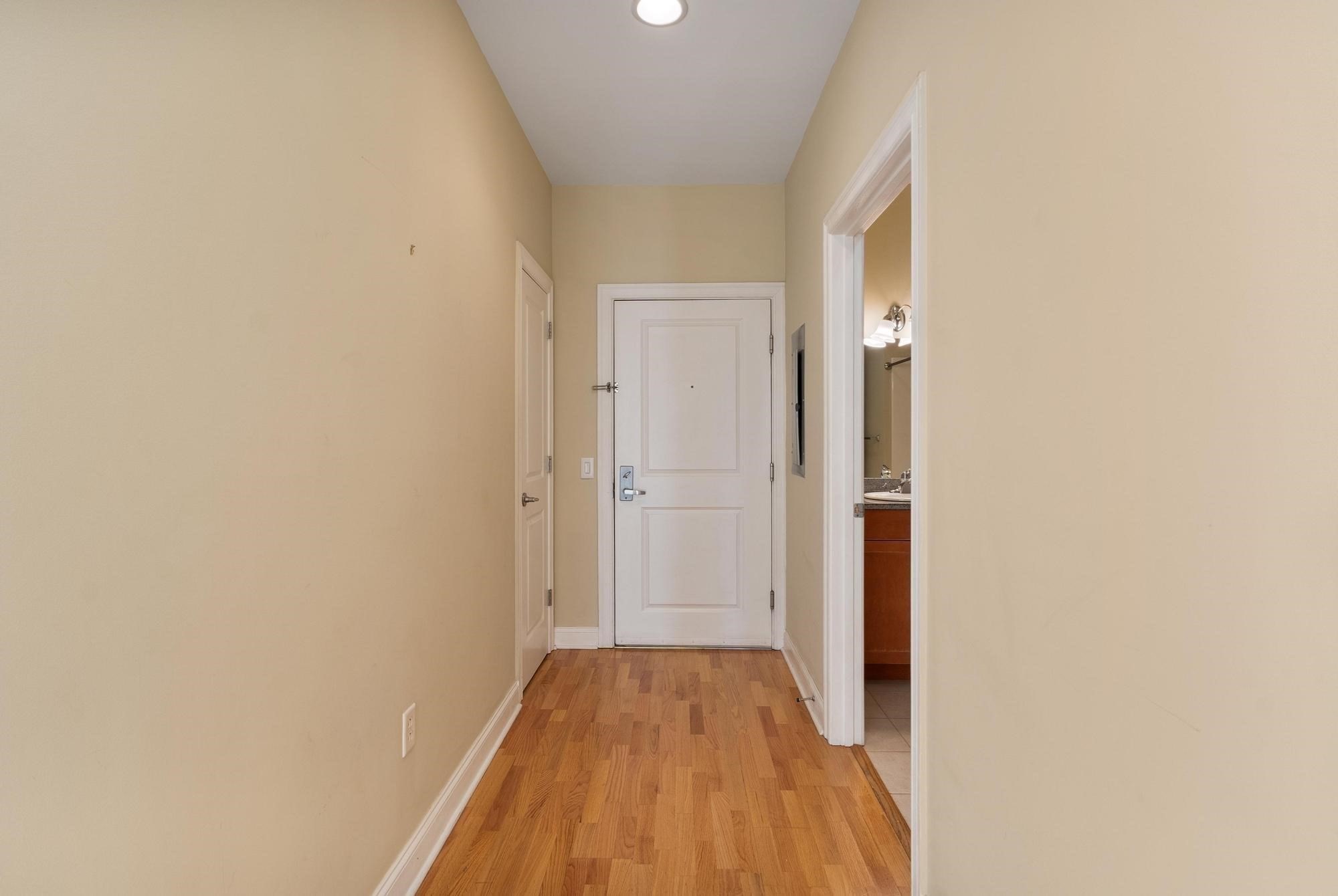 Hallway with hardwood floors leading to a white entry door, with a bathroom doorway on the right.
