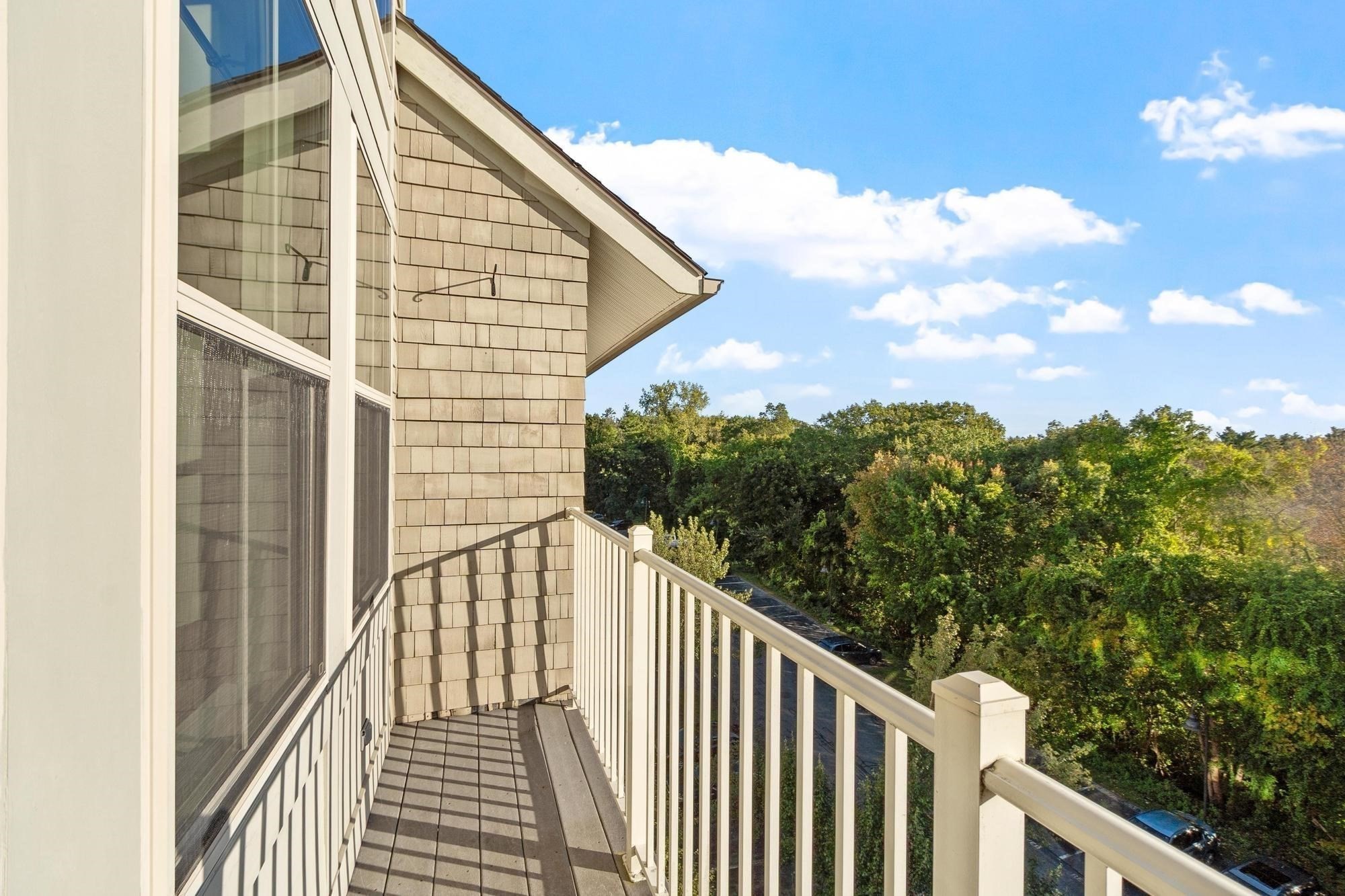 Second-story balcony with gray decking and white railing overlooking trees and a parking area.
