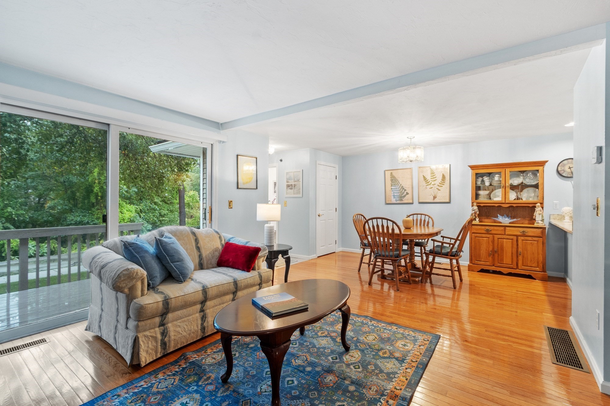 Living and dining area with hardwood floors, a blue patterned rug, and sliding doors overlooking trees.