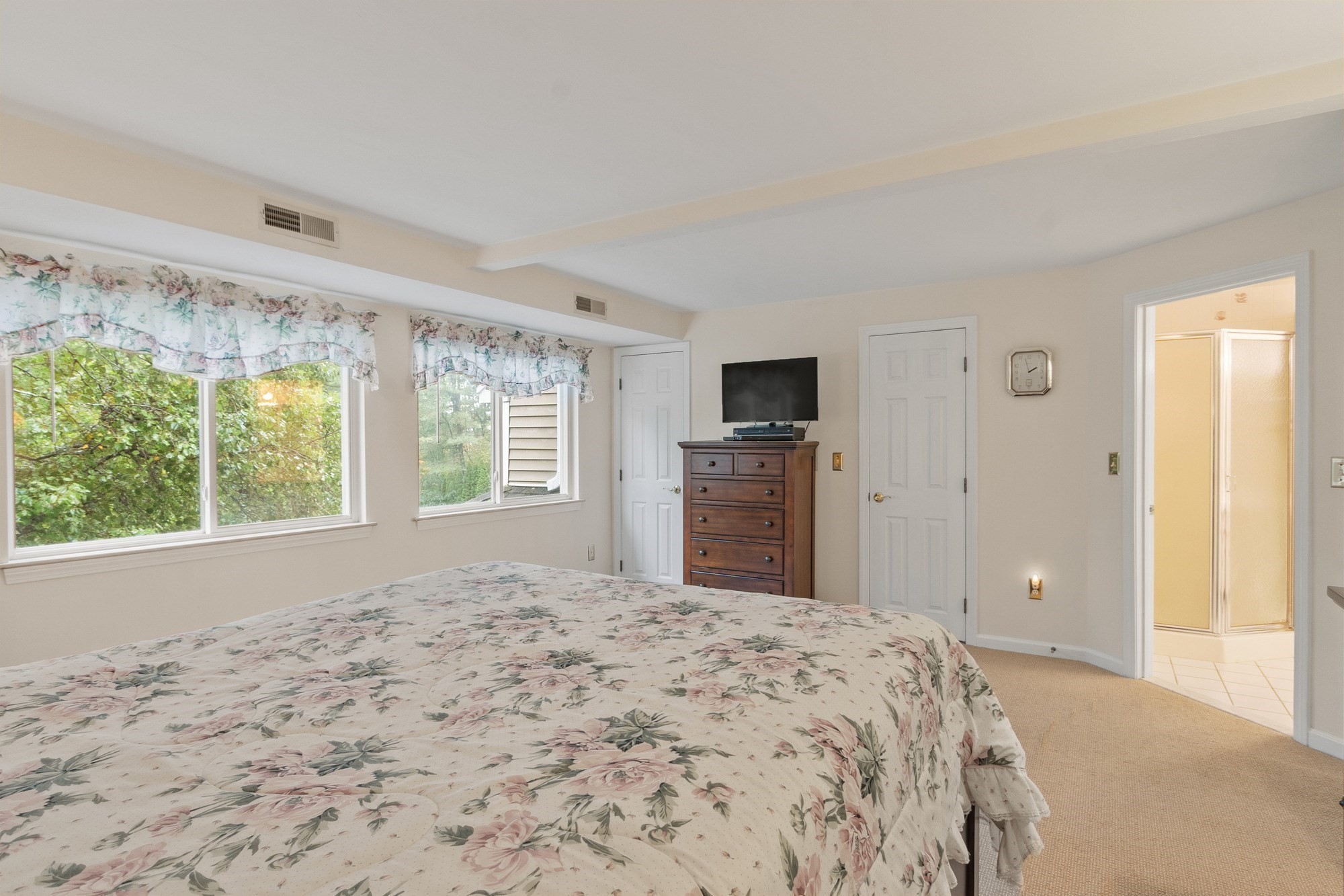 Bedroom with floral bedding, leafy views through two windows, and an open doorway to the bathroom.