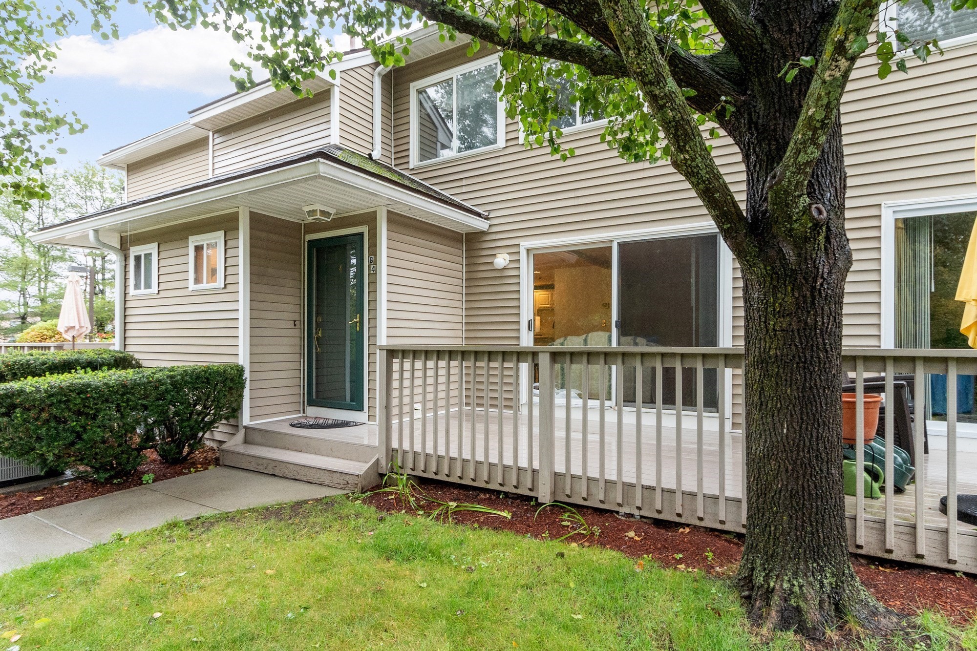 Beige two-story home with a covered entry porch and raised deck beneath a large shade tree.