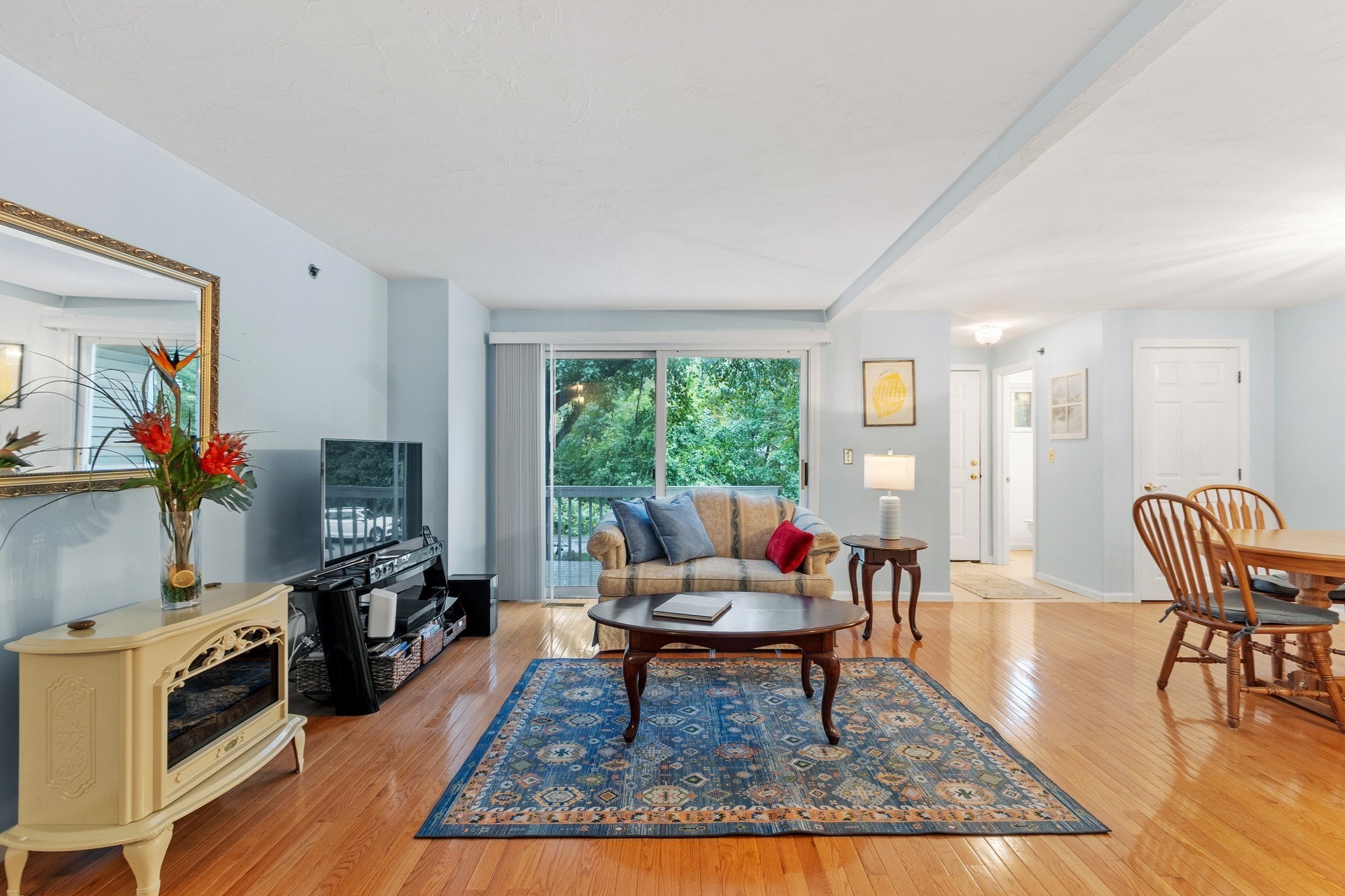 Furnished living room with hardwood floors, a blue patterned rug, and a balcony overlooking leafy trees.