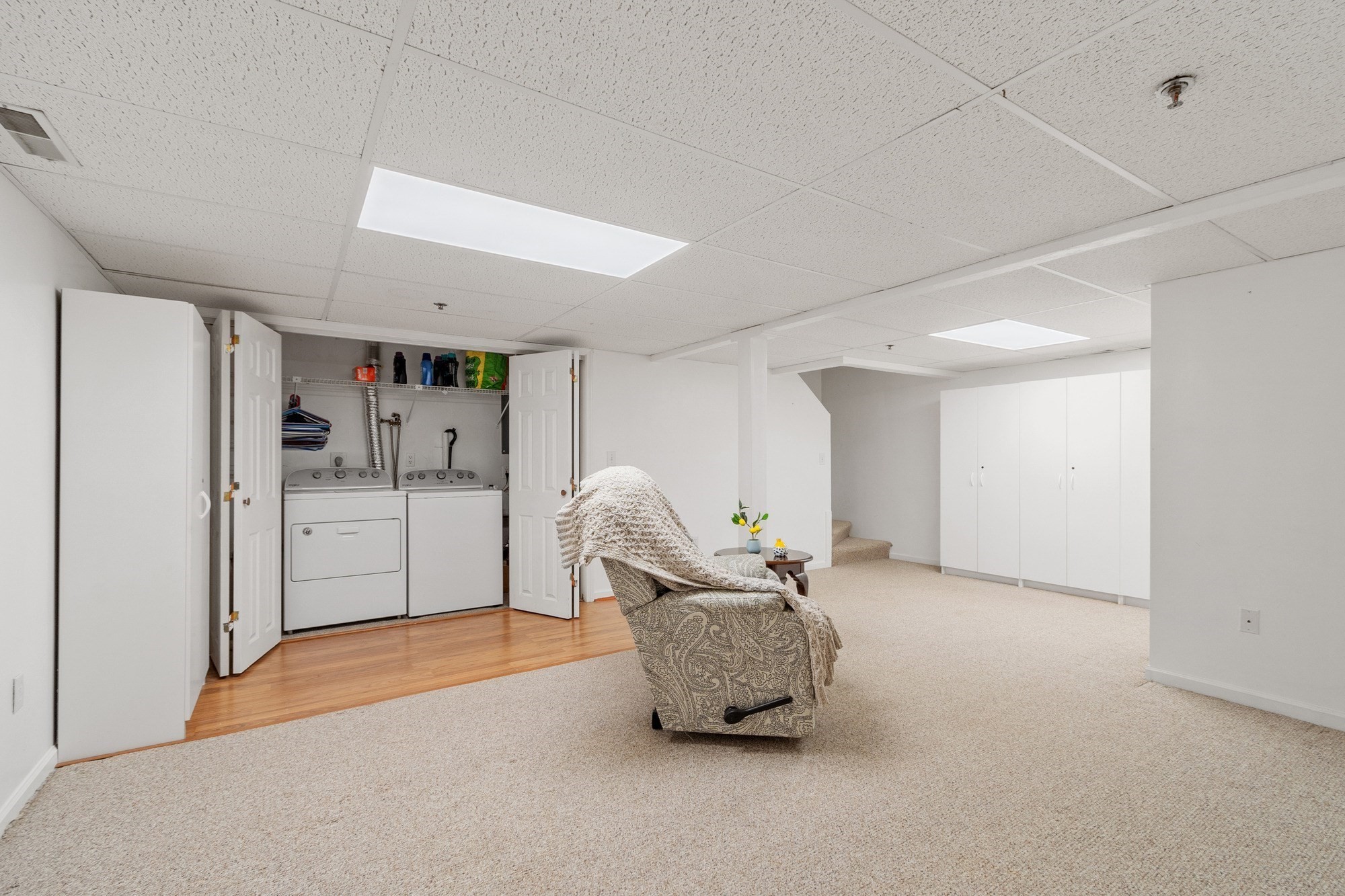 Finished basement with a laundry closet, patterned recliner, beige carpeting, and white storage cabinets.