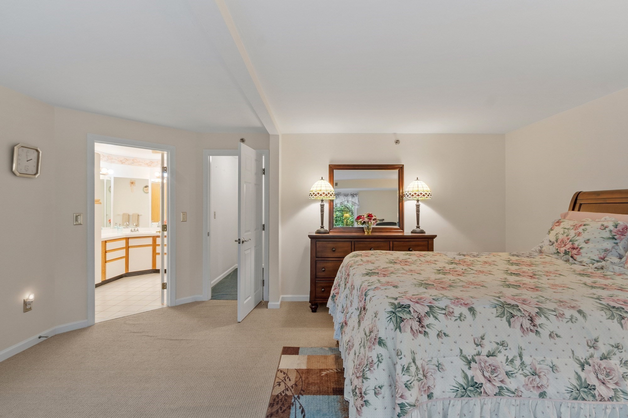 Bedroom with floral bedding, a wooden dresser and mirror, and an open doorway to a bright bathroom.