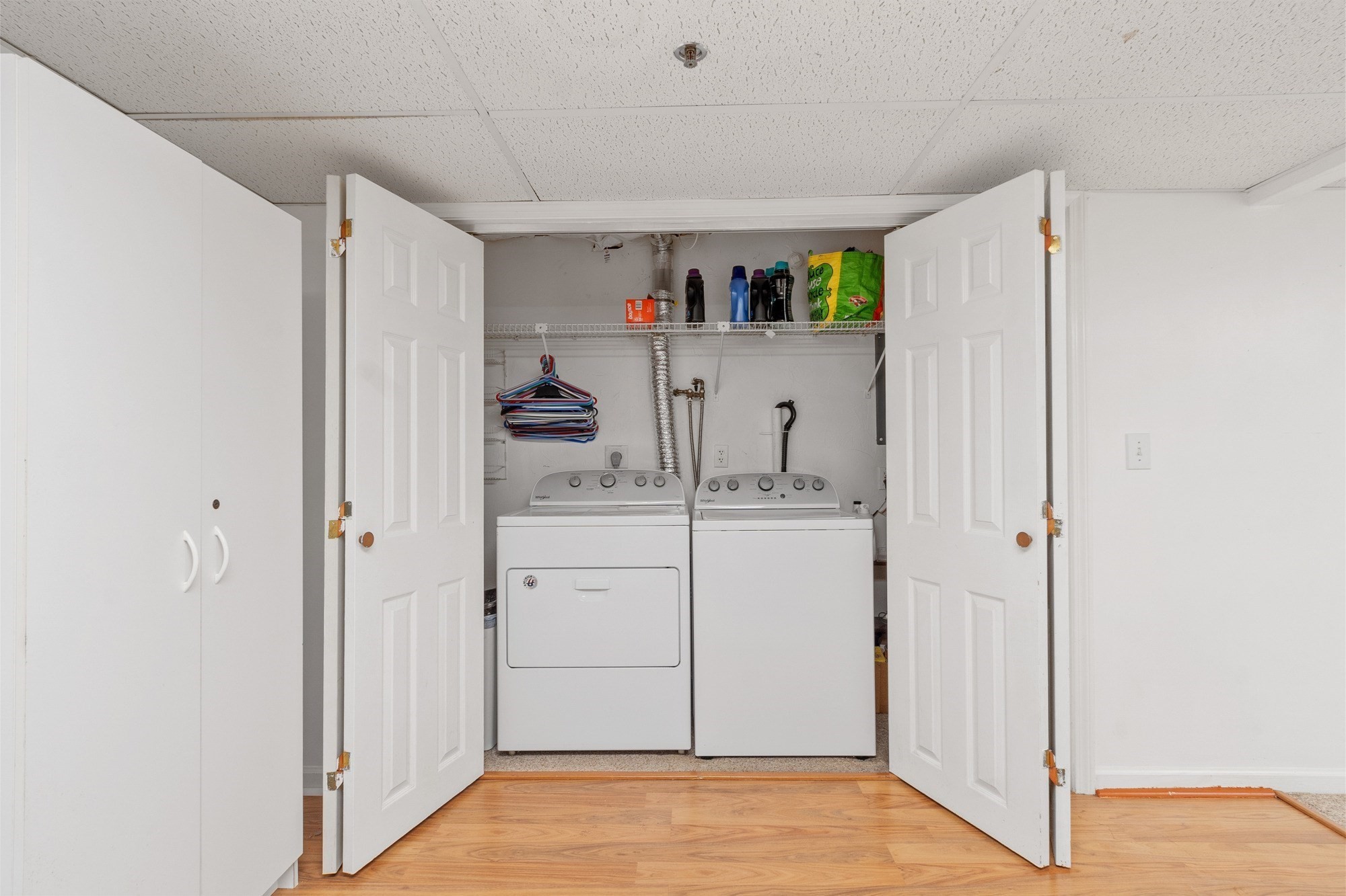 Laundry closet with open double doors, side-by-side washer and dryer, and a shelf of supplies.