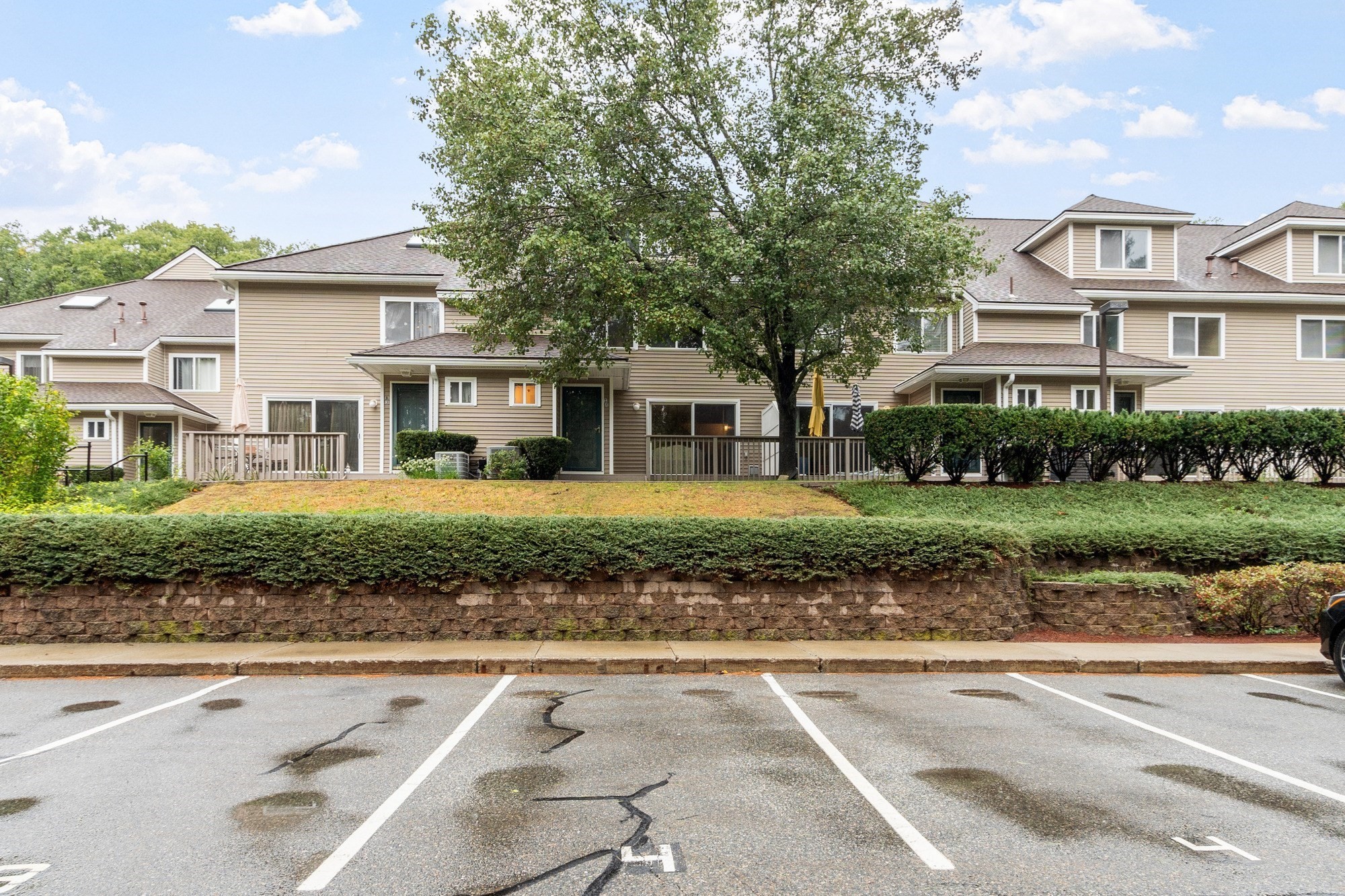 Beige multi-unit building with patio decks, a large tree, hedges, and parking spaces in front.