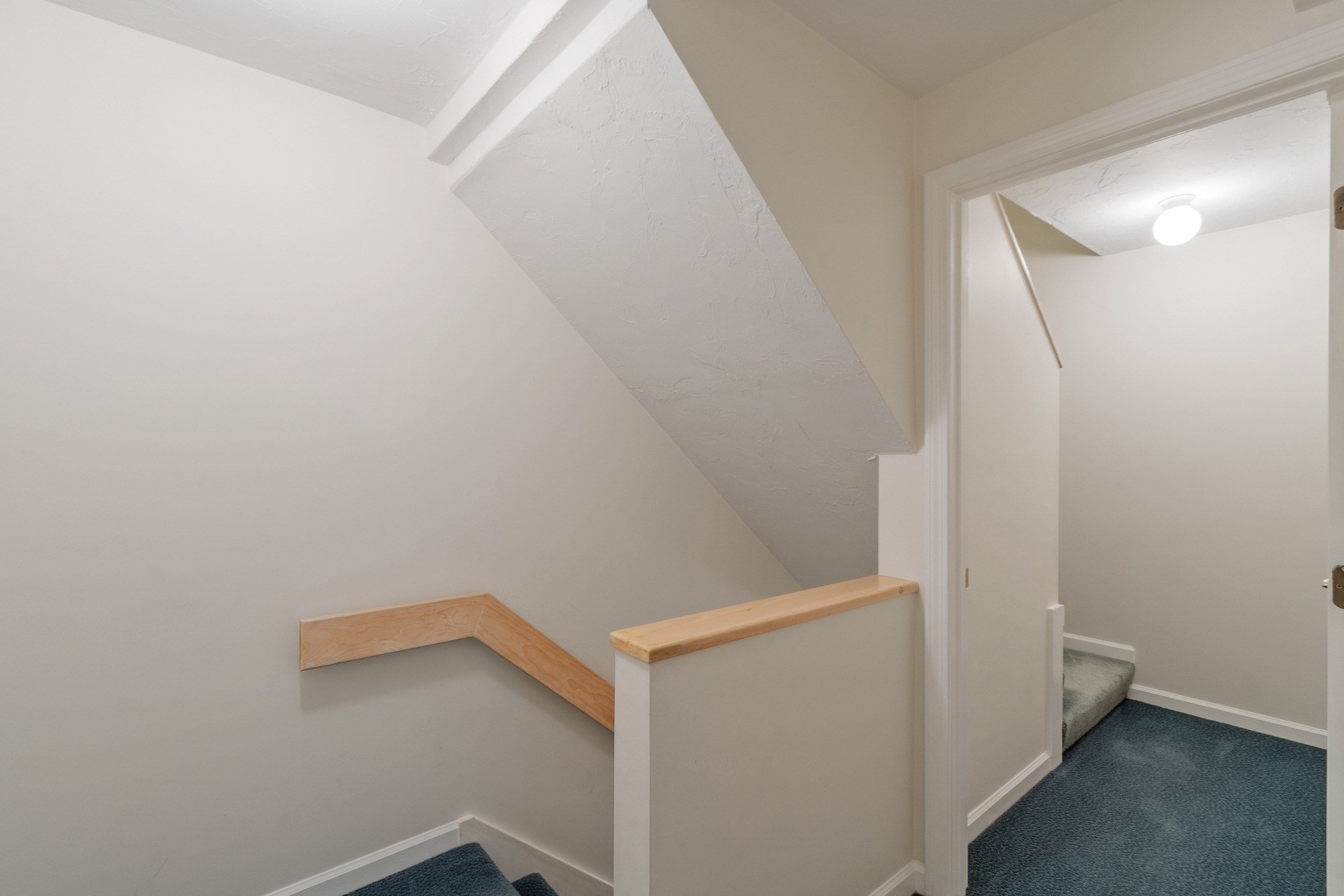Empty stairwell landing with blue carpet, a low sloped ceiling, and light wood handrails.