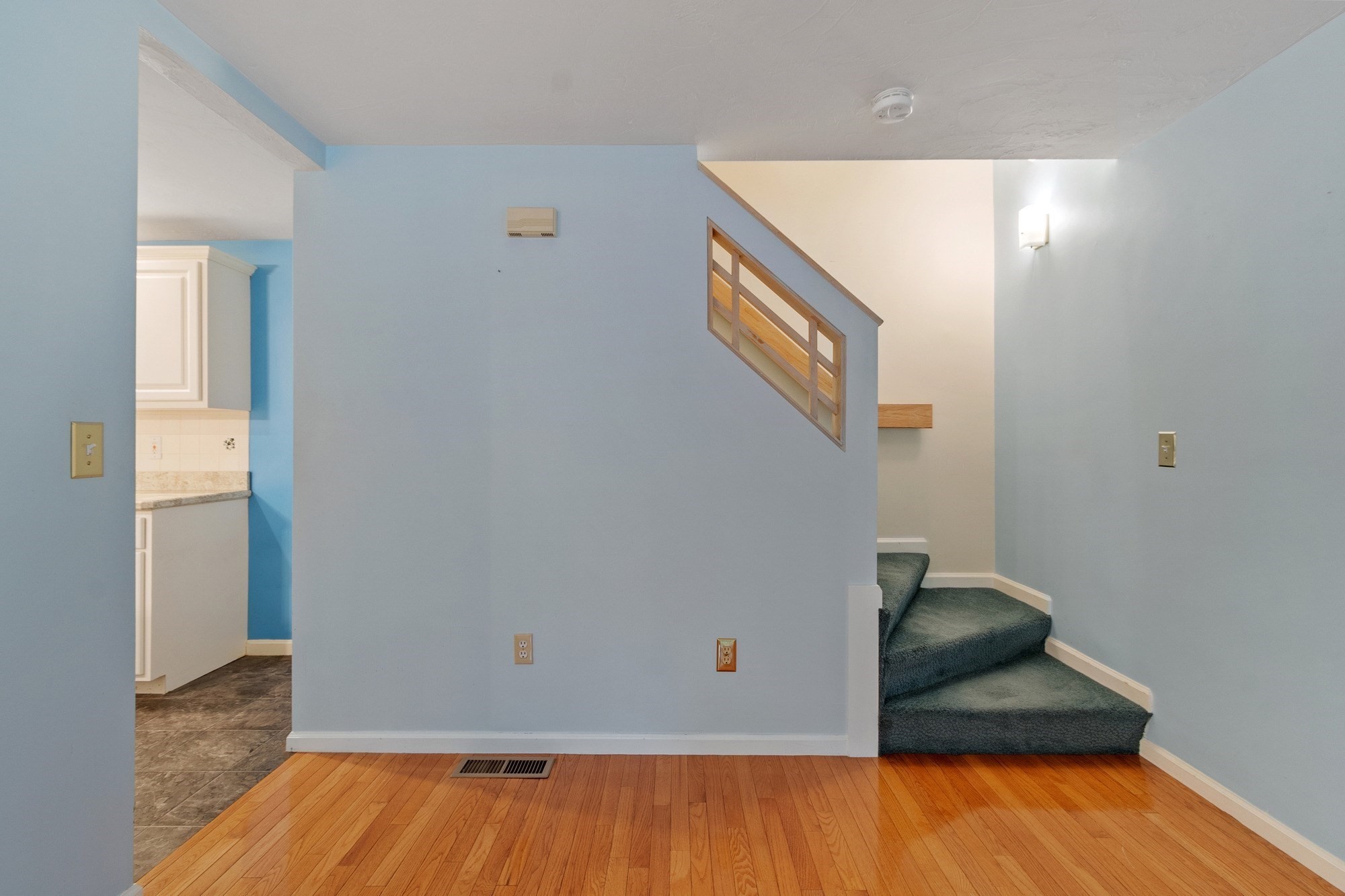 Vacant room with hardwood floors, blue walls, and carpeted stairs beside a kitchen opening.