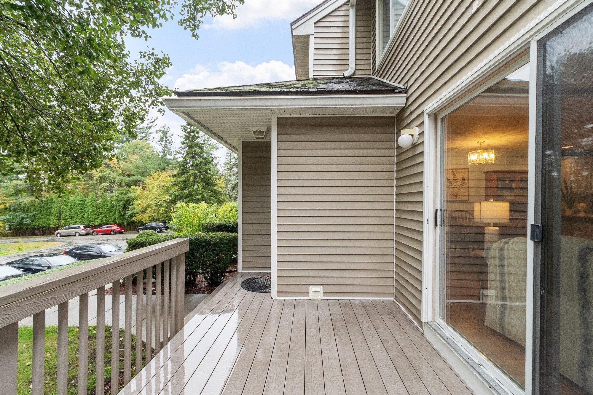 Raised deck with tan siding and sliding glass door overlooking trees and a parking area.