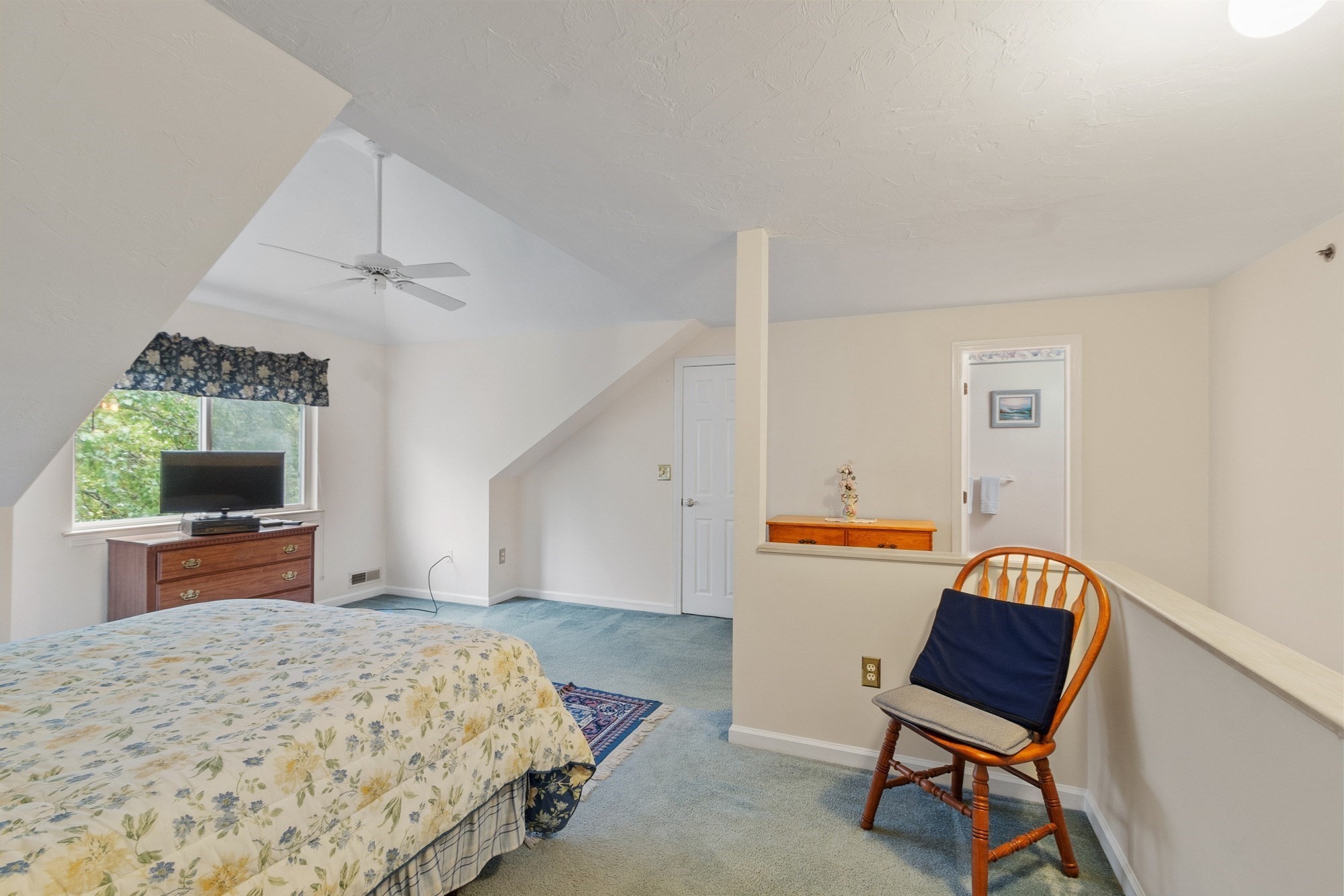Bedroom with a sloped ceiling, floral bedspread, and wooden chair beside the stair railing.