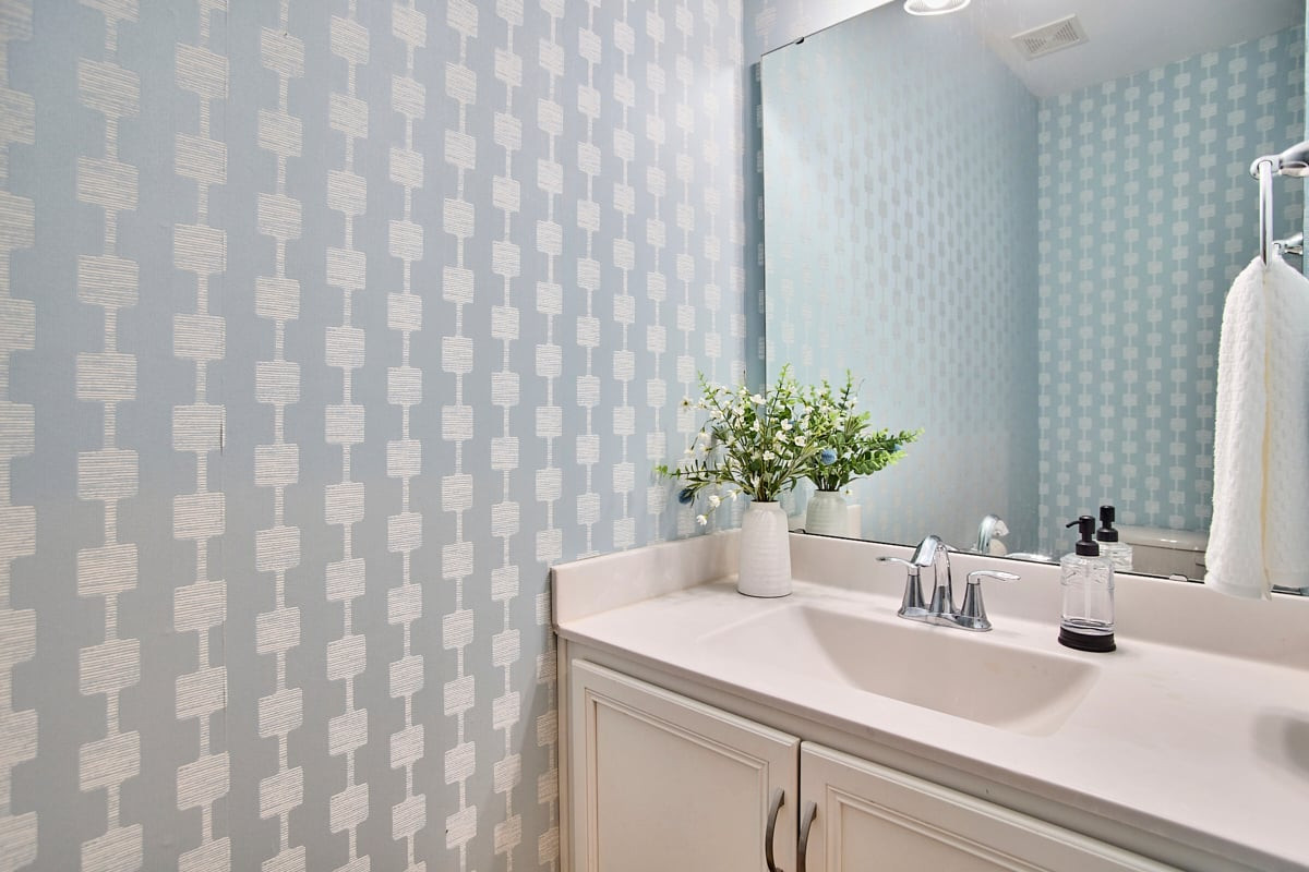 Bathroom vanity with a white sink, chrome faucets, mirror, and pale blue geometric wallpaper.