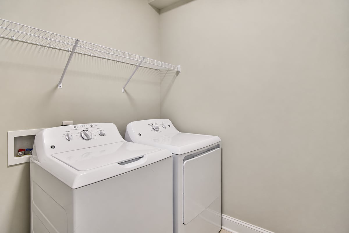 Laundry room with a top-load washer and dryer beneath a wire shelf.
