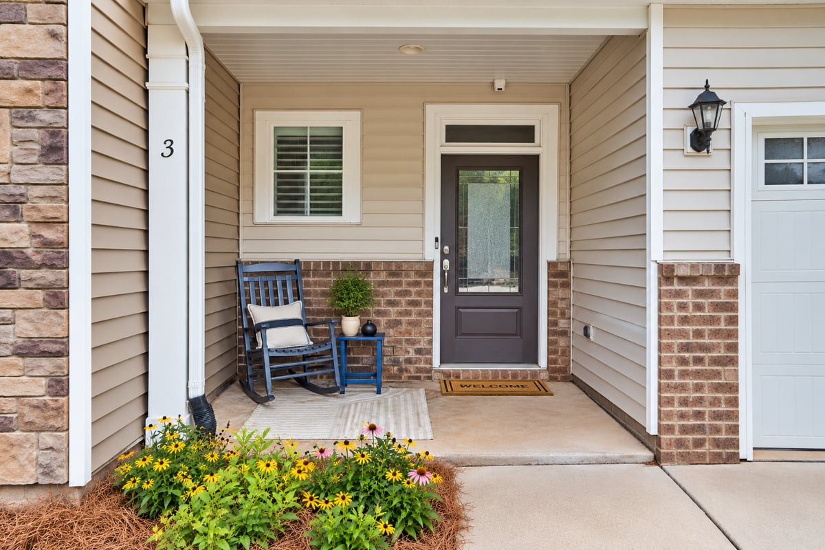 Home entry porch with a dark glass-paneled door, rocking chair, and yellow flowers along the walkway.