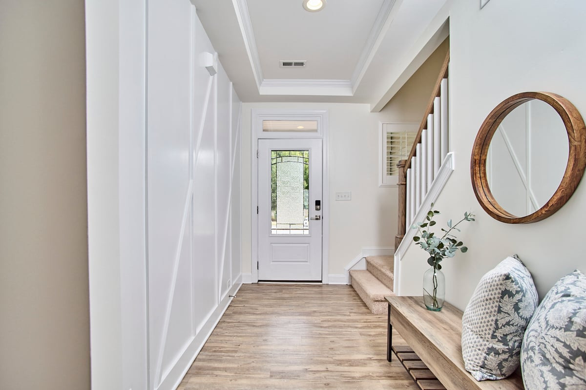 Bright foyer with a glass-paneled front door, carpeted stairs, and a wooden bench beneath a round mirror.