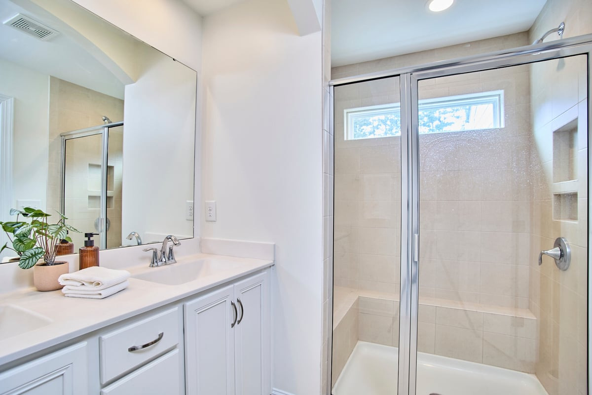 Bathroom with white vanity and glass-enclosed tiled shower featuring a built-in bench and high windows.