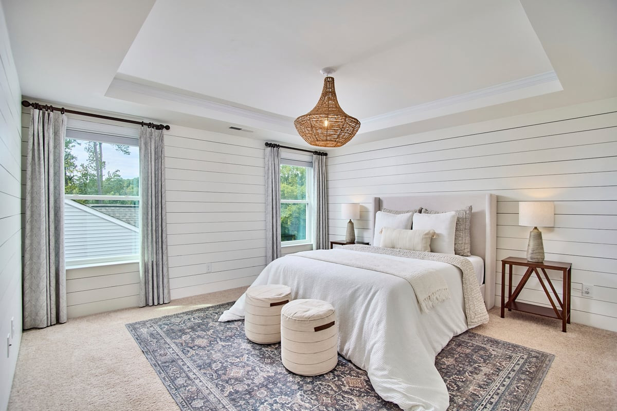 Staged bedroom with white bedding, shiplap walls, two curtained windows, and a woven pendant light.