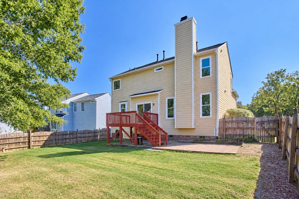Rear of a beige two-story home with a red deck, fenced lawn, and patio.