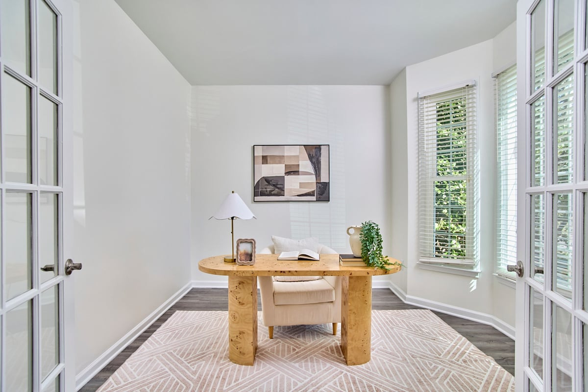 Bright home office with a wooden desk, cream chair, patterned rug, and tall windows.