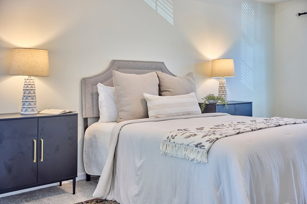 Staged bedroom with a gray nailhead headboard, white bedding, and blue bedside cabinets.