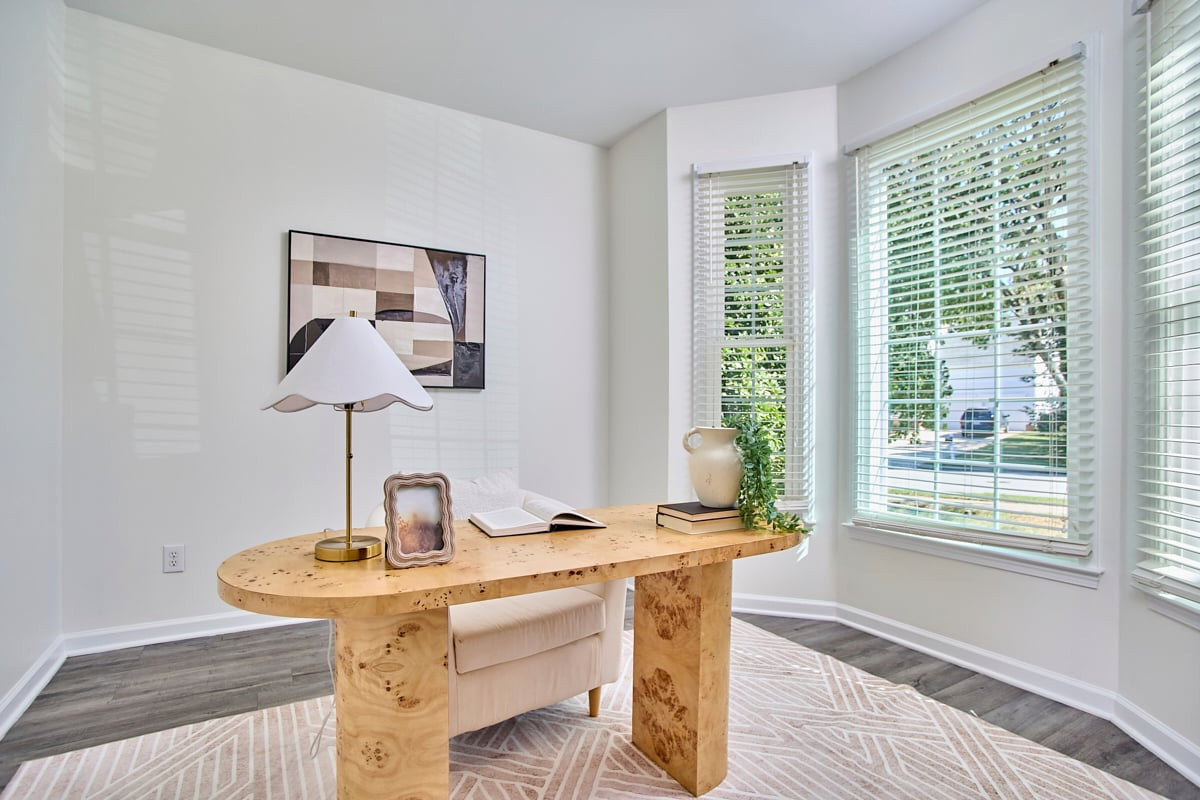 Bright home office with a sculptural wood desk, upholstered chair, and tall windows with blinds.