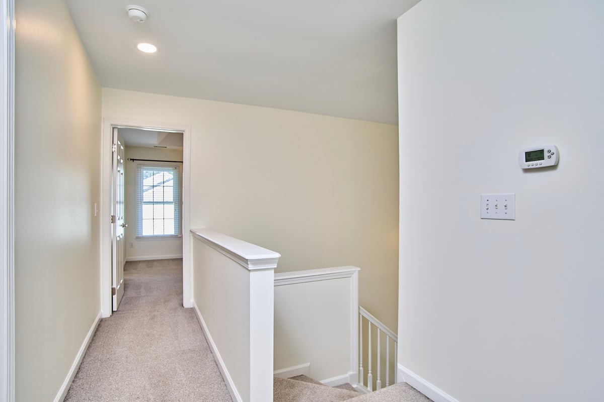 Carpeted upstairs landing with a stairwell, thermostat, and doorway to a bright room.