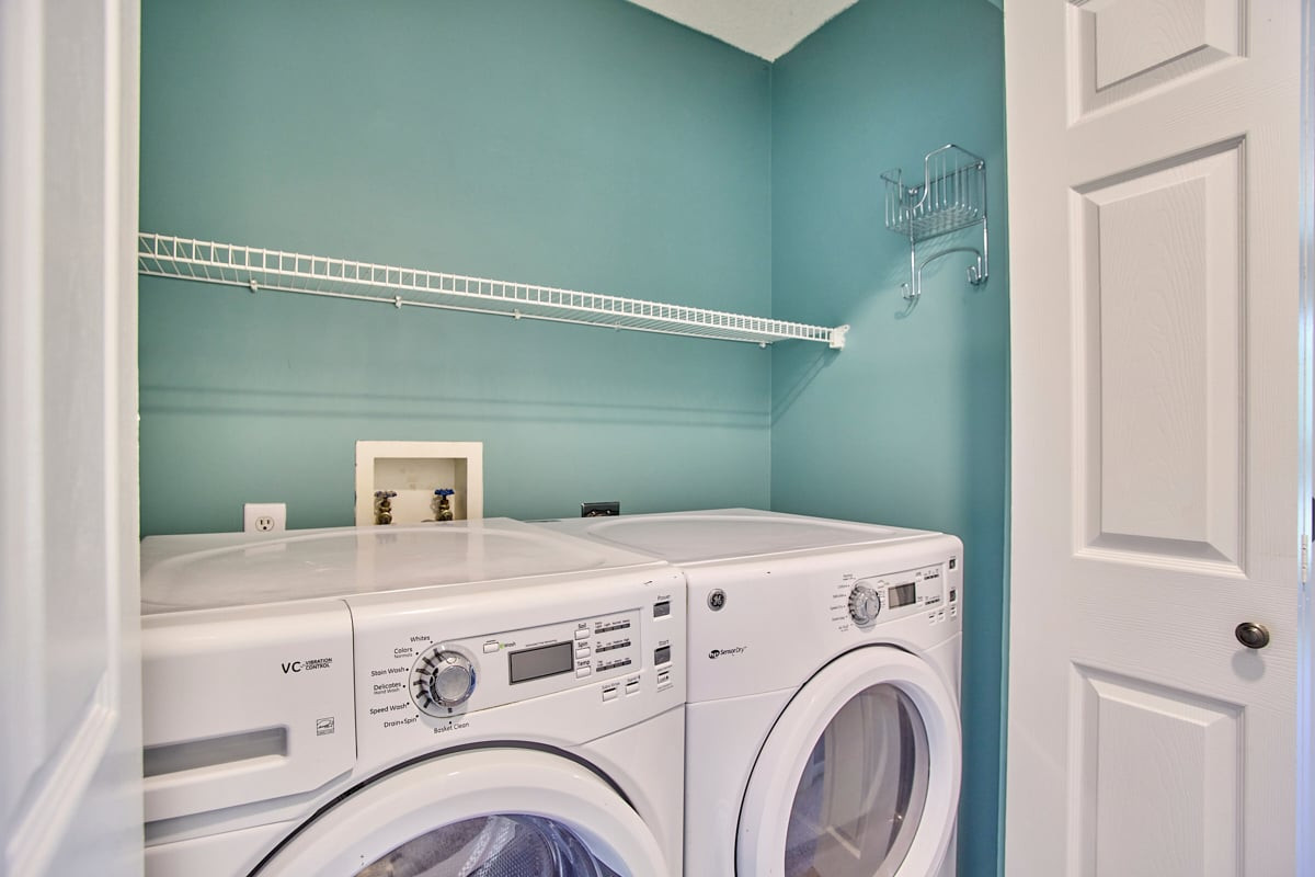 Laundry room with white front-loading washer and dryer beneath a wire shelf.