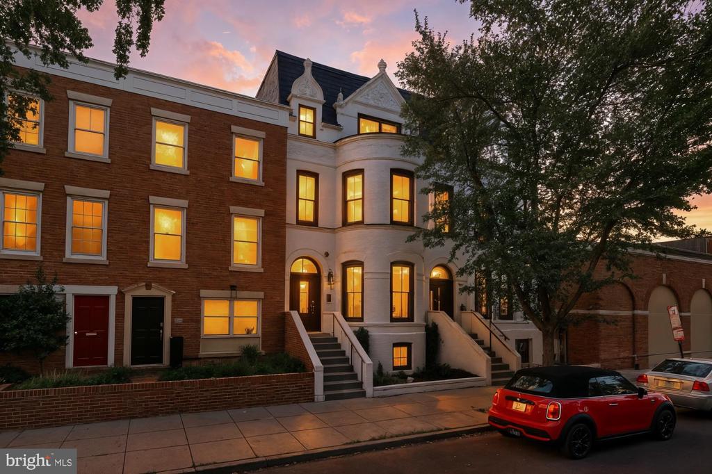 Brick and stone townhouses at dusk with glowing windows and a red car at the curb.