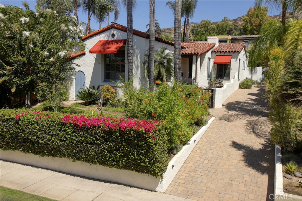 White stucco home with red tile roofs, flowering landscaping, tall palms, and a brick-paver driveway.