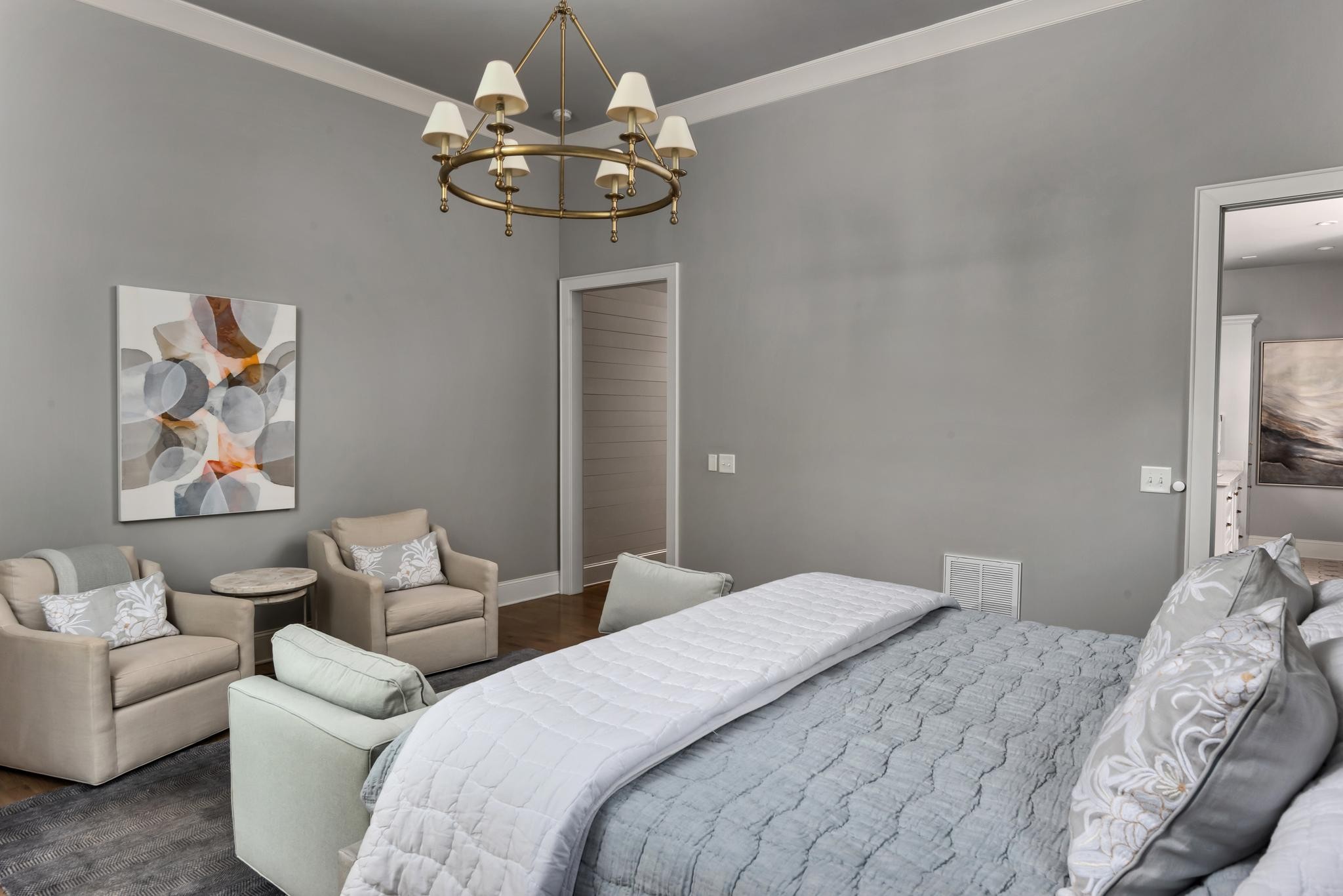Furnished bedroom with gray bedding, beige armchairs, abstract wall art, and a brass chandelier.