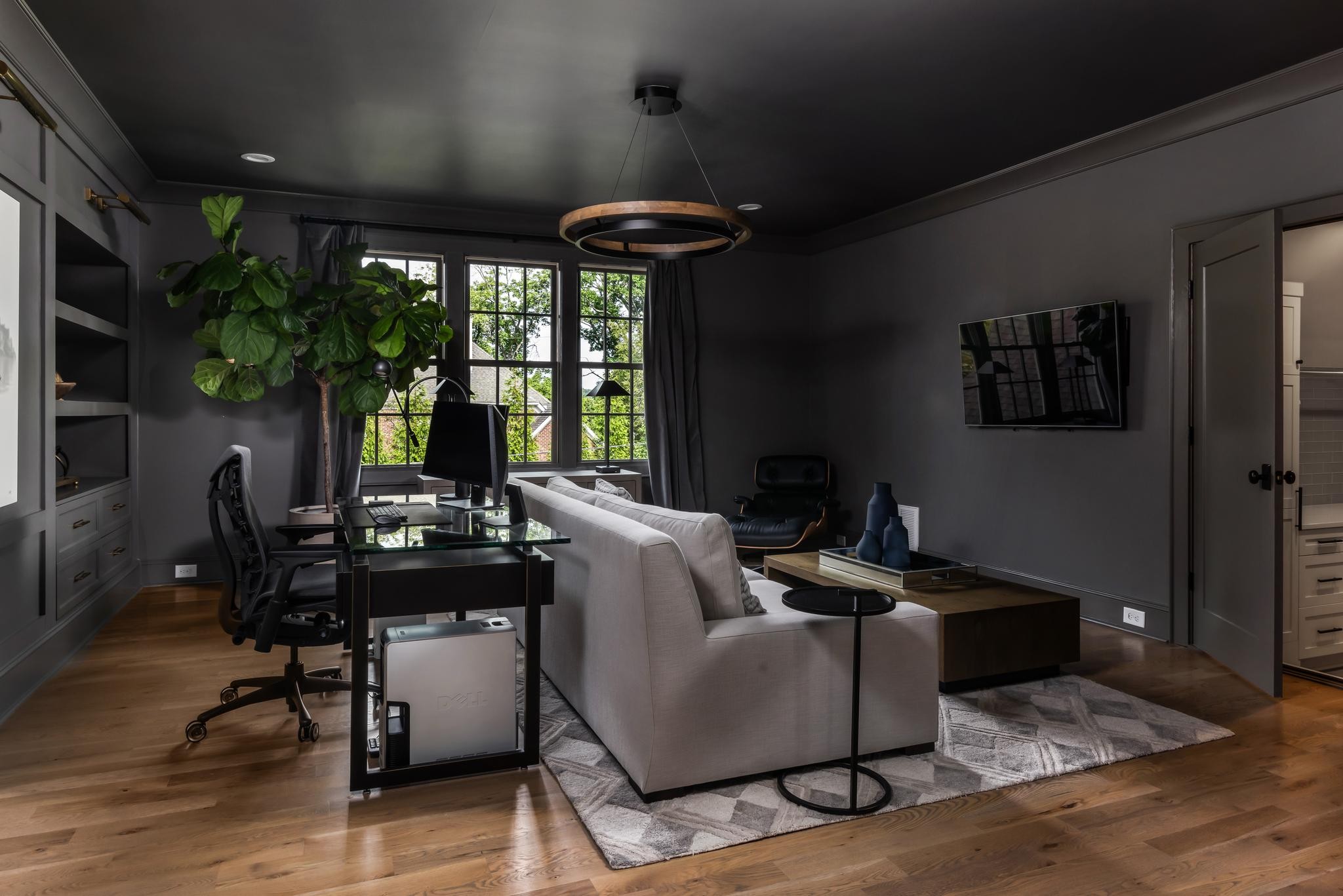 Dark living room with a glass desk, tall windows, leafy plant, and white sofa.