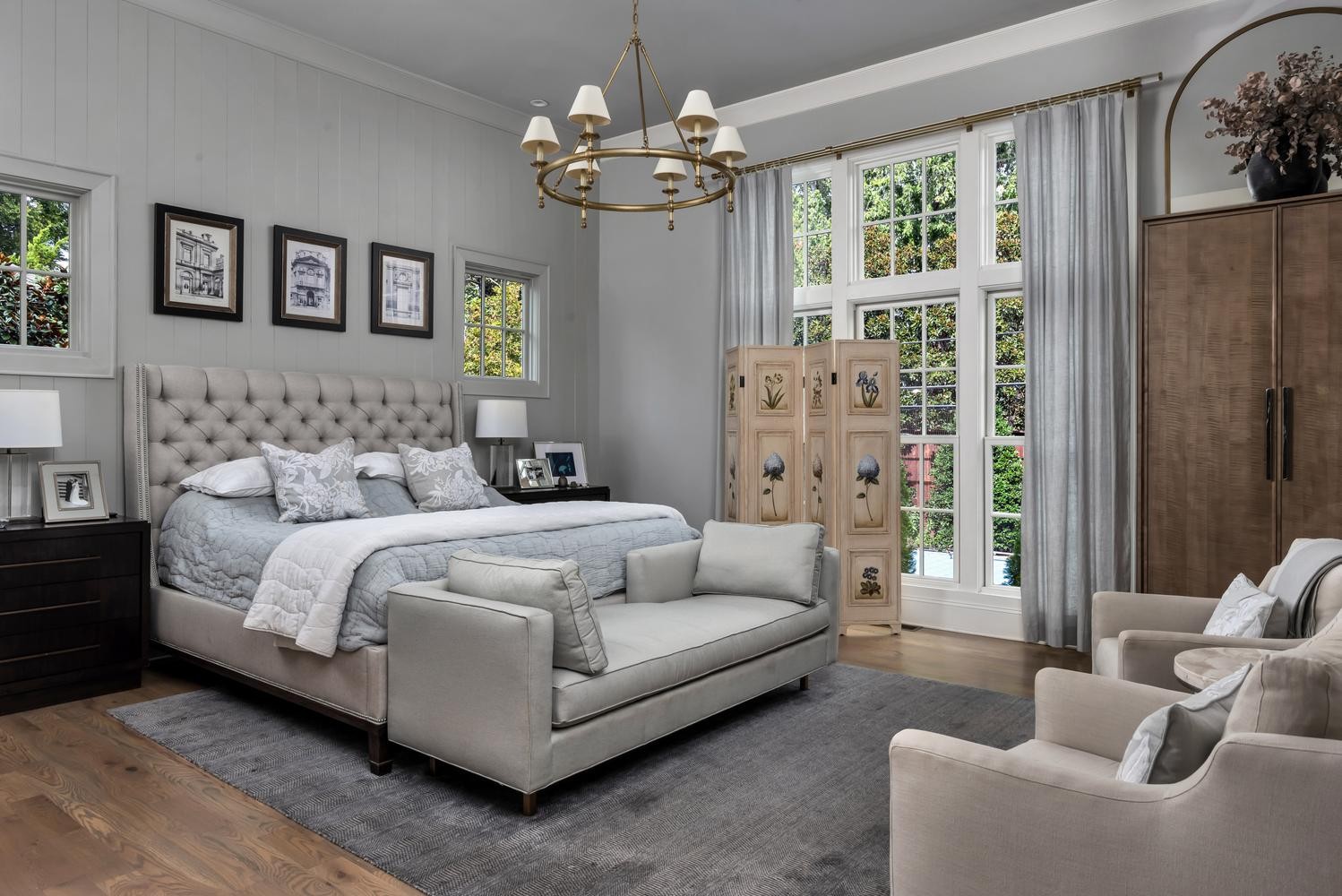 Staged bedroom with a tufted bed, upholstered bench, tall windows, and two armchairs.